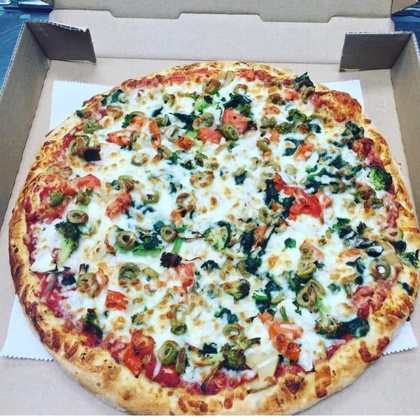 A Del Baggio Pizzeria veggie pizza with olives, broccoli, and tomatoes in a cardboard box.